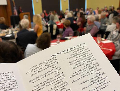 {A scholarship luncheon program in the foreground with a group of guests in the background}